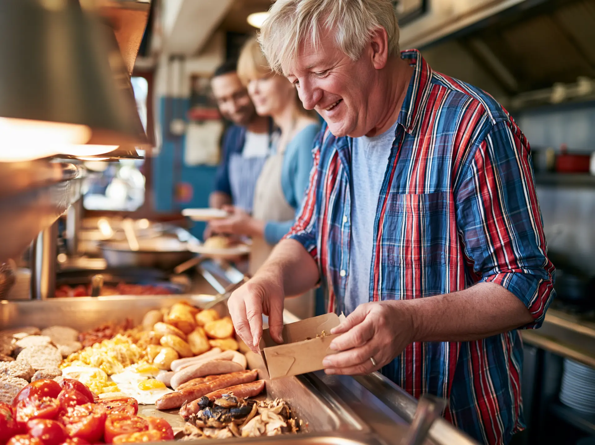 A man filling a takeaway box from a hot breakfast buffet line of sausages, eggs, tomatoes and mushrooms.