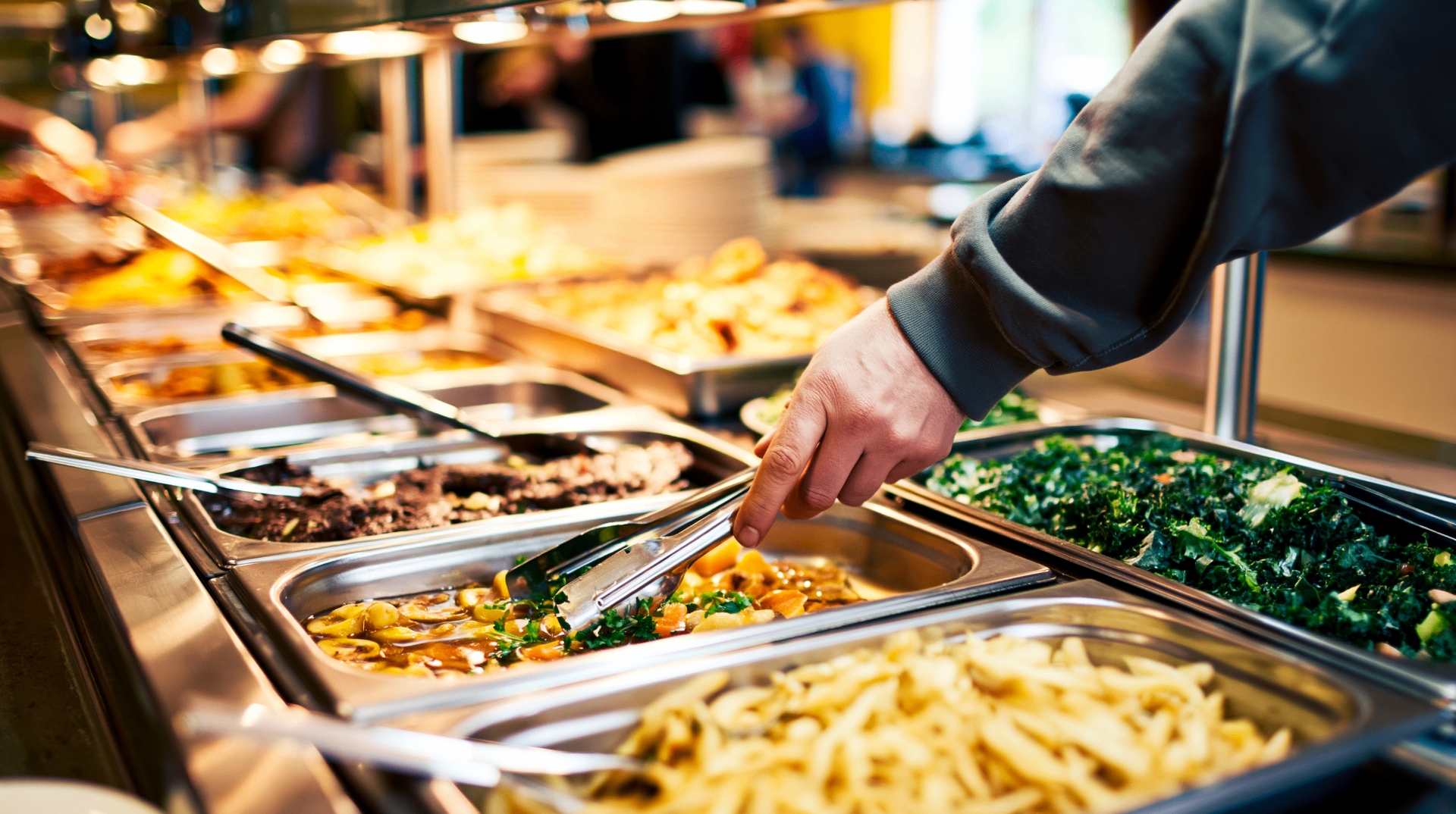 A buffet line of hot dishes — vegetables, curries and pasta — being served with tongs.